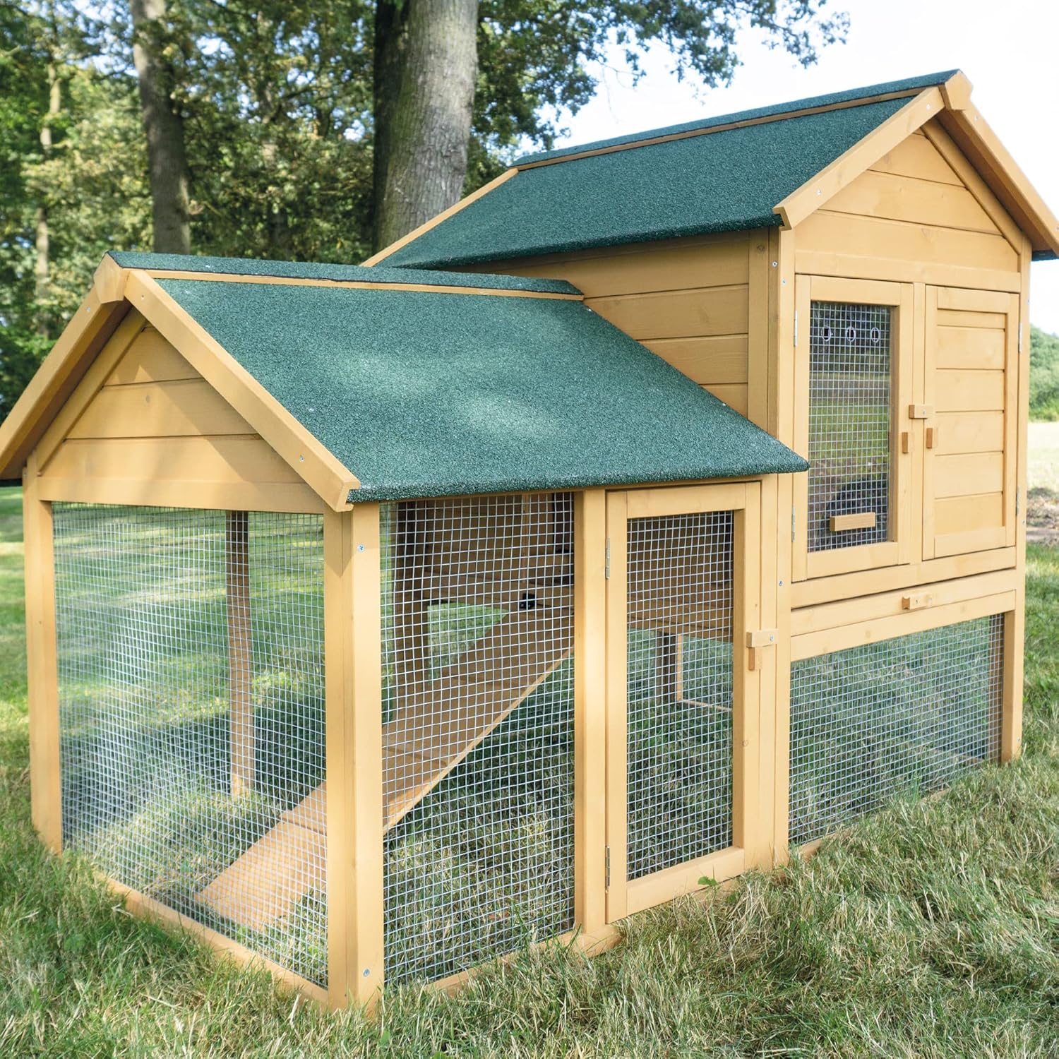 Ein Hühnerstall aus Holz mit grünem Dach, zweigeschossig gestaltet und mit einem Außengehege auf dem Rasen. Die Struktur umfasst eine Rampe, die den oberen und unteren Abschnitt verbindet, und befindet sich in einem schattigen, grasbewachsenen Bereich mit Bäumen im Hintergrund – perfekt für alle, die kaufen möchten.